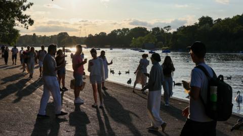 A crowd walks along the Serpentine in the evening light. It is sunny and lots of ducks, swans and pedalos are in the water.