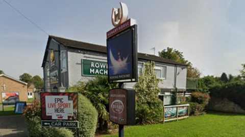 The Rowing Machine pub is a relatively compact building in front of a lawn on a street corner, with signs outside advertising the pub and TV sports coverage.