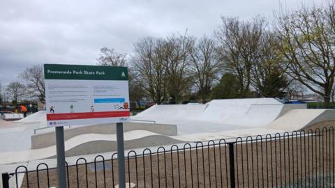 A concrete skate park. The concrete is white and cream. There are trees to the right. It is cloudy above. In the foreground is a dark-painted fence and a sign reading PROMENADE PARK SKATE PARK.