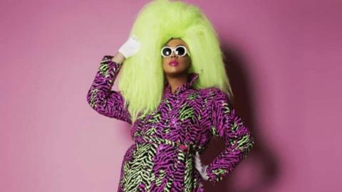 A black man with sunglasses in a large yellow wig and patterned pink and yellow dress against a pink background