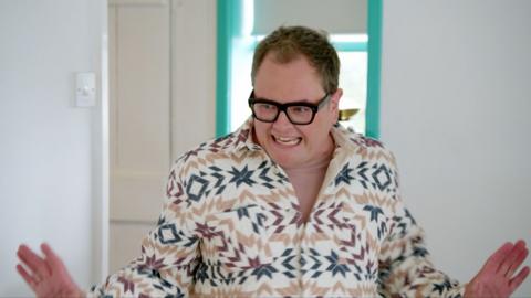 Alan Carr smiles with his hands outstretched. He wears black rimmed glasses and a patterned shirt