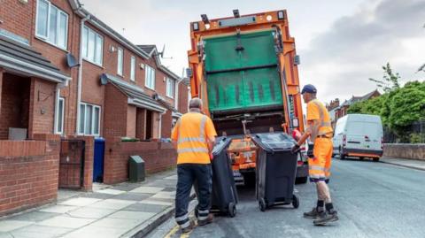 Two refuse collectors in orange high-vis. They are moving two black bins towards a truck.