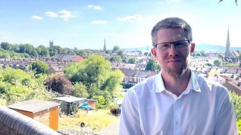 A man, who has short brown hair, a stubbly beard and is wearing glasses and a white shirt, is standing in front of a view of the city of Worcester with houses, steeples and greenery in the background. 