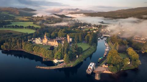 An aerial view of Fort Augustus and Caledonian Canal on the shores of Loch Ness. Mist covers parts of the villages and nearby hills.