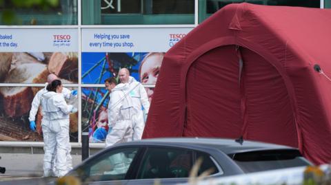 Four forensic workers dressed in white zip-up suits and wearing blue gloves stand outside a red tent outside Fairgreen Shopping Centre in Carlow. Behind the tent are large windows with Tesco signs.