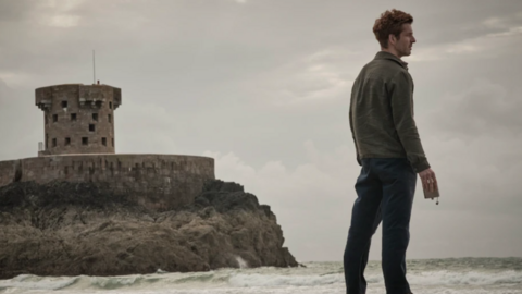 The main character of the Bergerac series standing by water looking out to sea. In the background is Jersey's Le Hocq tower.