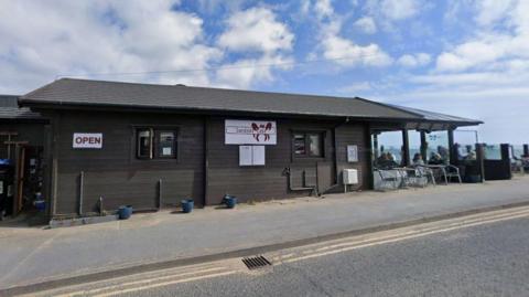 A small seaside café with dark wooden cladding and a sign reading “Sandside Café” sits beside a coastal road.