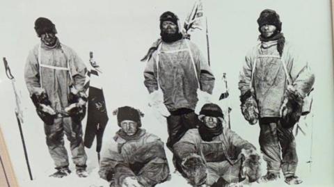 A black and white photo which has three men standing and two more sitting in front of them on the snow. They are wearing heavy clothing and hats and have ski poles. There is a Union Jack flag in the snow behind them.