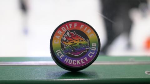 A hockey puck with the multi-coloured rainbow flag spread across it. There is also a dragon emblem on the puck.