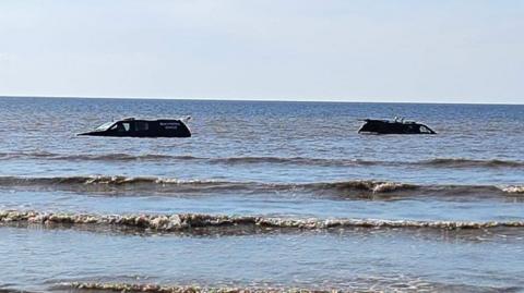 Two trucks are pictured in the sea, and small waves crashing into the shore.