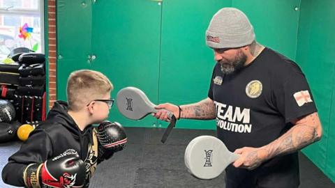 Oliver is punching two pads held by Neil Huntley in a gym. He is wearing a black jumper and has glasses. Neil is wearing a grey beanie and has a beard.