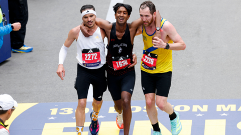 Three men wearing black shorts and different coloured running vests jog towards the finish line of the Boston Marathon with their arms around each other. Robson De Oliveira, who has short, dark hair and a moustasch and is wearing a white vest and a white headband. Ajay Haridasse has short, dark hair and a black vest. Aaron Beggs has short brown hair and a beard and is wearing a yellow vest with a blue stripe.