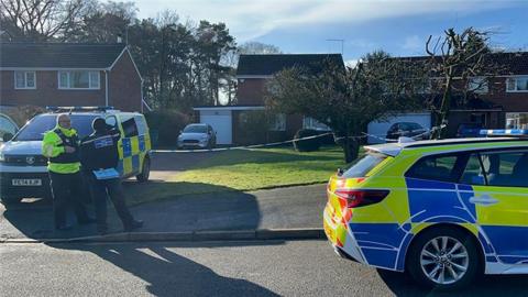 A police car and police van outside a house with white and blue police tape in front of the driveway. There is a car in the driveway and a tree in a small garden
