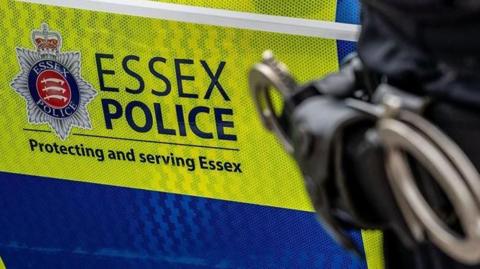 A green and blue Essex Police car with the hip of a police officer in front of it. That officer has a set of handcuffs on them.