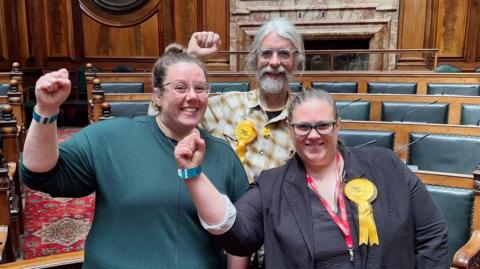 Two women and a man wearing yellow rossettes hold their clenched right fists in the air in celebration. They are standing in a wood paneled room.
