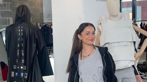 Young woman with long dark hair and a dark jacket, standing next to a model wearing a long cloak