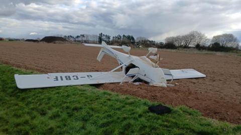 A small white aircraft which is upside down and has crashed onto a patch of brown grass on an airfield. There is damage to the front of the aircraft. The plane's nose is wedged into the ground and the doors to the aircraft are open.