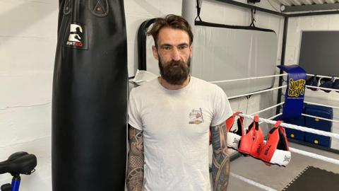 Jamie Parker wearing a white t-shirt with a logo on the right. He has tattoos on both arms and a beard. There is a gym ring in the background with white ropes and red and white boxing gloves. He is stood next to a large black punching bag.