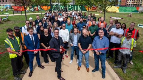 A man wearing glasses and a blue blazer holds giant scissors, cutting a red ribbon, in front of large crowd comprising people in work clothing and others dressed casually, in a large green space with a sign reading "Aire Park" in the background.