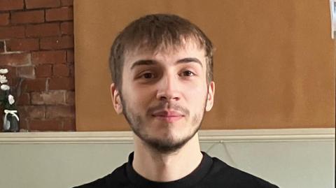 A young man with short brown hair and beard wearing a black T shirt smiles at the camera