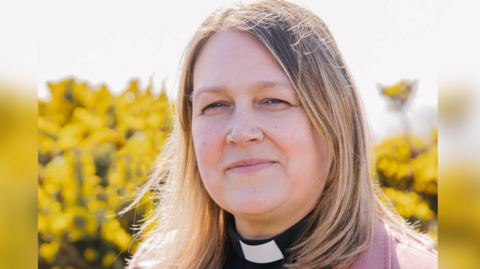 Close up of the Rev Leah Vasey-Saunders against a flowery background. She has shoulder-length blonde hair and is wearing a dog collar.