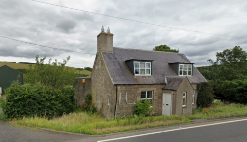 A stone cottage next to a road. The sky is grey and the house is surrounded by a lot of overgrown shrubs and grass