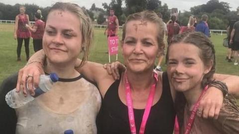 Three women pose for a photo having done a muddy run. The mother is in the middle wearing a black top and medal with a pink lanyard. She has her arms around her two daughters. The one on the right is a teenager, with brown plaited hair and wearing a brown top and a medal. On the left is an older woman in her twenties, wearing a light top and holding a water bottle.
