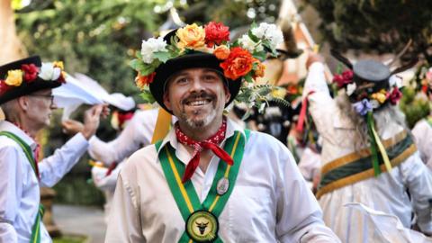 Duncan Bhaskaran Brown wearing Morris dancing attire, including a white shirt and a cross of green and yellow fabric over his shirt, a red tie around his neck, a black top hat with flowers on it.