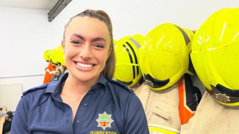 Millie wears her hair up in a pony tail and is dressed in her firefighter's uniform. There are helmuts and uniforms on pegs on the wall behind her.