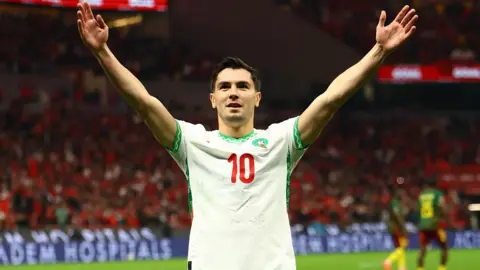 Brahim Diaz, wearing a white Morocco shirt with green trim and a number 10 in red on the chest, holds his arms aloft in celebration