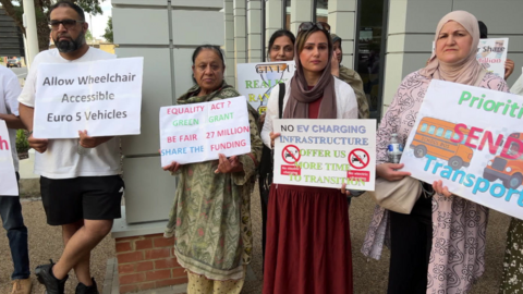 A group of people - four women and one man - holding placards. Nazneen, who is wearing a red dress and a poster that says "No EV charging infrastructure, offer us more time to transition".