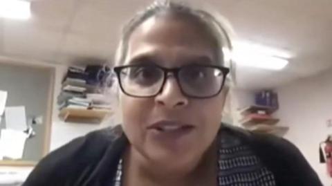 Kuldeep Stohr looks at the camera during an NHS video she is taking part in. She is wearing a black and white blouse and a black cardigan. She has black, plastic-framed glasses on and greying hair which is tied back. She is sitting in an office with folders packed onto shelves in the background and some papers pinned to a board.