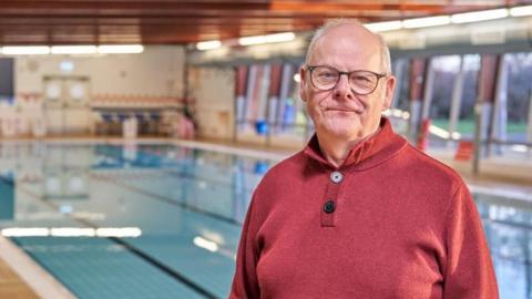 A man wearing glasses and a red top stands next to a swimming pool. 