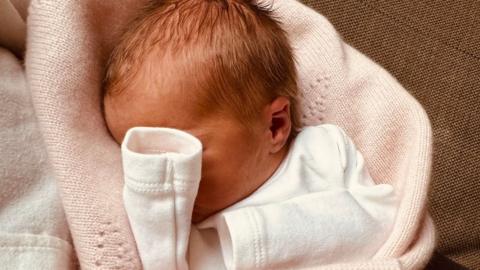 A newborn baby girl is pictured in a close-up photograph showing most of her head, with her hand covering much of her face, apparently fast asleep. Her hands are hidden in her sleeves and she is wrapped in a pale pink blanket.