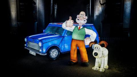 Three lanterns from Longleat's Festival of Light annual event. One is a blue car and the other two depict Wallace and Gromit - a stop-motion character wearing a white shirt, red tie, green jumper vest and orange trousers. He has one hand on his hip and a thumb up. Next to the Wallace lantern is Gromit, a cream dog with brown ears and a big black nose. 