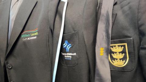 Three grey school blazers hanging on a rail. One has William Lovell CofE Academy logo, another has Thomas Middlecott Academy and the third has a logo consisting of three golden crowns