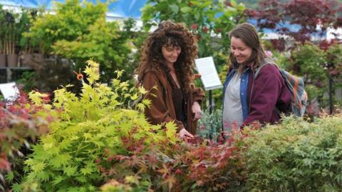 Two women are standing among rows of shrubs and are smiling. The woman on the left, who has long hair and is wearing a brown jacket, is touching some leaves. 