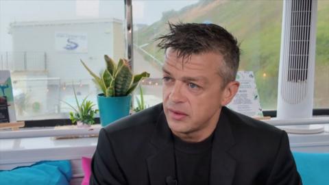 Former PC Lorne Castle sits giving a TV interview. He has short dark brown hair with grey flecks. He is wearing black blazer with a black round neck t shirt and he is sat on a blue sofa.