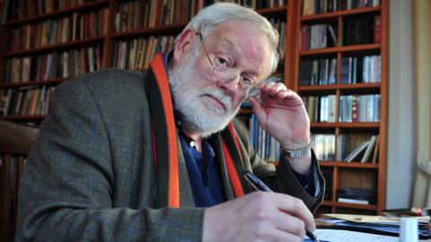 Michael Longley staring into the camera over the lense of his gold glasses. He is pulling the glasses down with his left. He has white hair and a white beard with gold glasses. He is wearing a dark tartan patterned coat and a red, orange and dark green striped scarf. He is writing with a fountain pen at a desk with bookcases full of books behind him.