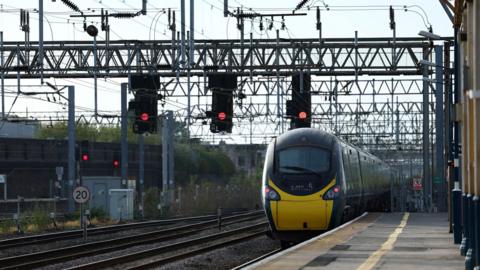 A train pulls into Crewe railway station. There are red lights on overhead gantries and a white circular speed sign with a black 20 in the middle with a red border. The platform has a yellow line running along it, and there are pillars on the far right hand side of the image.