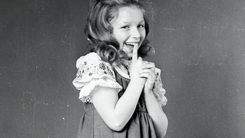 A black and white photo of a young Lena Zavaroni smiling with her finger at her mouth. She is wearing a pinafore and a floral trimmed blouse with puff sleeves.