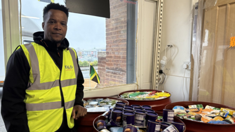 A man with short black hair stands besides four large bins that are filled with food. He is wearing a black hoodie and a high viz jacket.