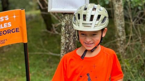 Louis wearing a bright orange t-shirt and a helmet. He is holding an orange flag.