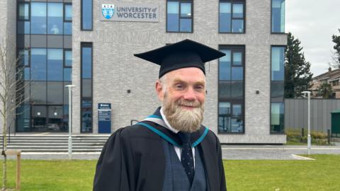 A man with a bushy light/dark grey beard stands smiling at the camera. He is wearing a black mortarboard and a mid-blue suit with a dark blue tie under a black graduation gown. Behind him is a grey brick building with a sign reading: "University of Worcester".