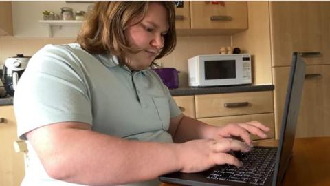 Kye Vallance sits at his laptop typing from his kitchen