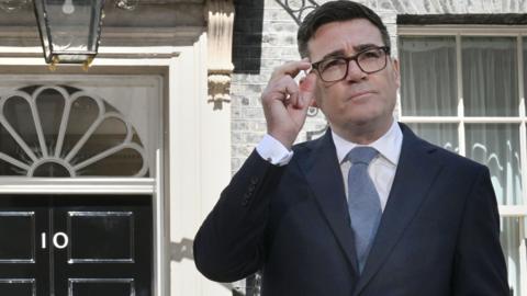 Andy Burnham, wearing a dark suit and glasses, standing outside the front door of 10 Downing Street