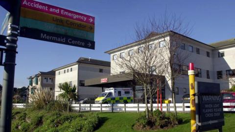 Several buildings at the Royal Cornwall Hospital in Truro on a sunny day. An ambulance is parked outside one of the buildings. Three cars are also parked outside. A multi-coloured sign pointing towards different areas of the site including the A&E department and Mermaid Centre is in the foreground of the photo. Trees and shrubs are on a grassy area in front of the buildings.