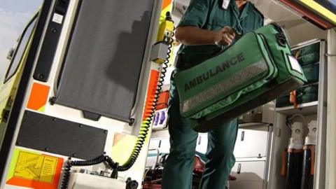 Man wearing green top and trousers stepping down from the back of an ambulance. He is carrying a green kit back with the word "AMBULANCE" written on it. The inside of the ambulance can be partially seen.