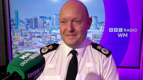 Mr Guildford is sitting in ˿ WM studio with a cityscape background and a green microphone in front of him. He is wearing a police uniform of a white shirt and black tie. He has a bald head.