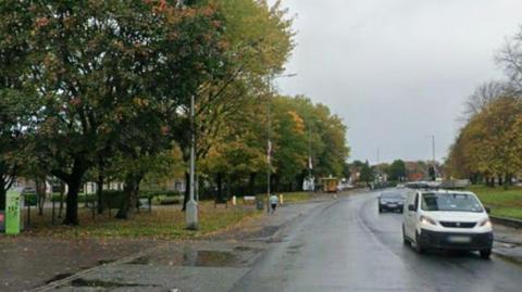 Warrington Road at the junction with Chatsworth Road, Rainhill on a rainy day in the autumn. There are trees either side of the road and a white van is seen driving towards the camera with a car in the background. There are brown fallen leaves on either side of the carriageway